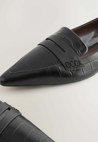 Schwarze Lederlofers mit spitz zulaufender Zehenpartie und strukturiertem Krokodilmuster. Mit einem Riemen und gestickten Details entlang der Kanten akzentuiert.