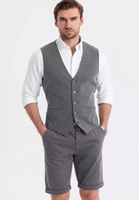 Gilet grigio a fantasia con cinque bottoni abbinato a pantaloni corti grigi. Camicia bianca con bottoni sotto, e orli arrotolati sui pantaloni.