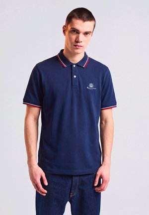 Junger Mann, der ein dunkelblaues Poloshirt mit rotem und weißem Besatz an Kragen und Ärmeln trägt, steht vor einem schlichten hellen Hintergrund.