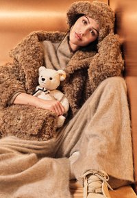 Abrigo y sombrero color tostado, hechos de material texturizado. Conjunto gris claro, sosteniendo un oso de peluche blanco. Ambiente suave y acogedor con colores cálidos.