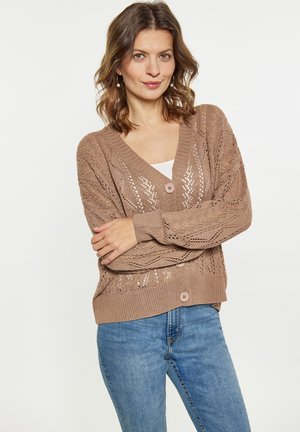 Frau mit schulterlangem, welligem Haar trägt eine braune gestrickte Strickjacke über einem weißen Top und blauen Jeans, steht mit verschränkten Armen vor schlichtem Hintergrund.