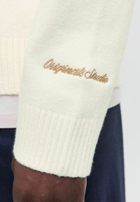 Close-up of dark-skinned hand wearing a cream knitted sweater with beige "Originals Studio" embroidery on the cuff.