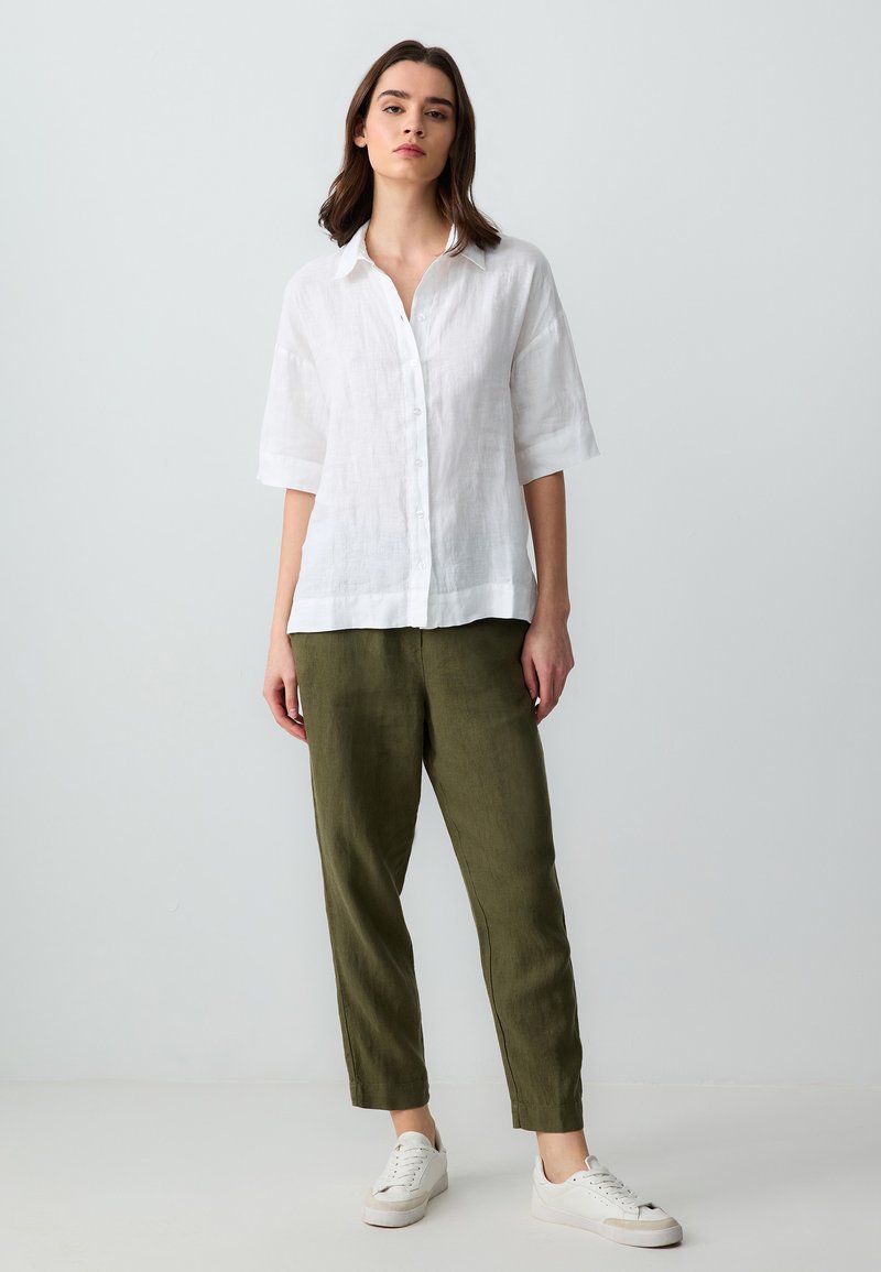 Femme debout portant une chemise blanche ample à boutons, un pantalon vert olive et des baskets blanches, sur un fond clair uni.