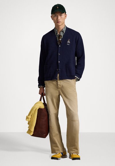 Cárdigan de rayas en color azul marino con un logo, camisa de cuadros, pantalones beige, suéter amarillo colgado sobre un bolso de cuero marrón, y zapatillas amarillas con detalles en negro.