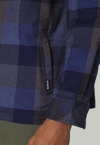 Zbliżenie na rękaw w ciemnoniebiesko-brązową kraciastą koszulę z mankietem i małą etykieta Barbour blisko nadgarstka.