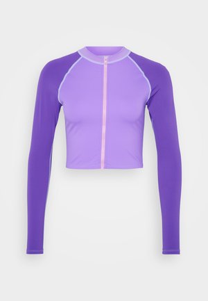 Vijolična skrajšana majica z dolgimi rokavi, ki ima zadrgo spredaj in kontrastne svetlo modre šive. Gladek, raztegljiv material.