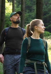 To vandrere i skoven: den ene har en sort langærmet skjorte på, den anden en grøn sweater. Begge har rygsække og ser opad.