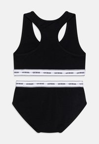 Ensemble de débardeur noir et de culotte avec une ceinture élastique blanche arborant le logo "GUESS" ; texture lisse et design dos nageur pour le confort.