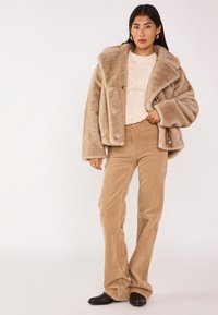 Cappotto in pelliccia sintetica beige con ampi risvolti, abbinato a un maglione a maglia crema e pantaloni a zampa d'elefante in corduroy beige. Stivali neri a punta completano il look.