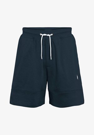 Man in een wit T-shirt met korte mouwen, marineblauwe shorts, witte sportsokken en witte sneakers, staand tegen een effen achtergrond.