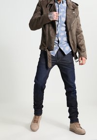 Hnědá semišová motocyklová bunda, modrá vzorovaná košile, tmavé džíny, béžové tenisky. Bunda má zipy a detail s postrann�ím páskem.