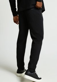 Zwarte joggingbroek van een zachte, elastische stof met een taps toelopende pasvorm, zijzakken met rits en elastische boorden, gecombineerd met zwarte sneakers.