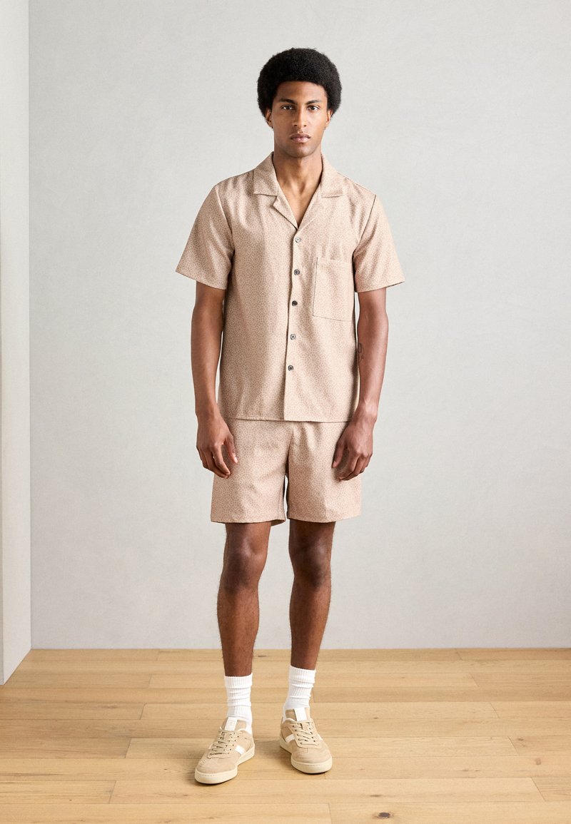 Camisa de manga corta color beige y pantalones cortos a juego con un patrón sutil. La tela parece ligera; cuenta con un bolsillo y cierre de botón. Se lleva con zapatillas.