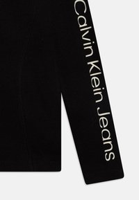Black ribbed fabric with a fitted design. One sleeve features the text "Calvin Klein Jeans" in white. Simple construction with no additional patterns.