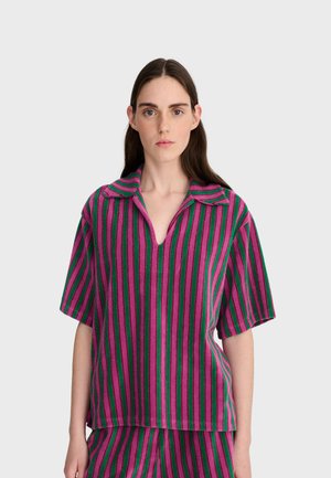 Femme aux longs cheveux foncés portant une chemise à manches courtes à rayures verticales violettes et vertes avec col, assortie à un bas, debout devant un fond uni.