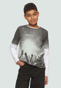 Grafik-T-Shirt mit einer Menschenmenge in Graustufen und erhobenen Händen. Darüber getragen wird ein weißes Langarmshirt. Dunkle Hose.