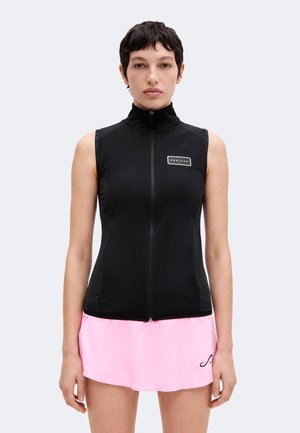 Personne aux cheveux courts et foncés portant un haut noir sans manches avec fermeture éclair et une jupe courte rose, faisant face à l'avant sur un fond uni.