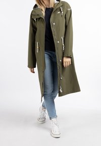 Veste imperméable vert olive avec capuche, dotée de cordons de serrage, de boutons et de poches latérales, associée à un jean bleu clair et des baskets blanches.