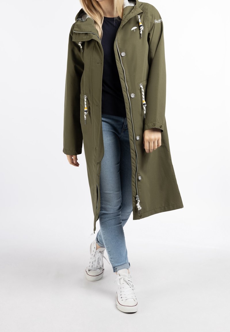 Veste imperméable vert olive avec capuche, dotée de cordons de serrage, de boutons et de poches latérales, associée à un jean bleu clair et des baskets blanches.