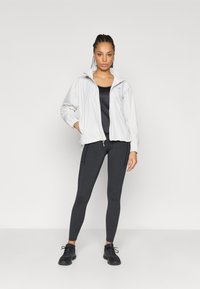 Jaqueta leve branca com fecho éclair, combinada com uma camisola preta sem mangas e leggings pretas. Tecido texturizado, ajuste justo e punhos elásticos. Ténis pretos.