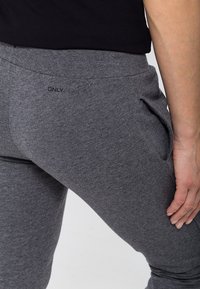 Zbliżenie osoby noszącej szare spodnie dresowe z boczną kieszenią i widocznym logo "ONLY" w pobliżu pasa, z ręką spoczywającą przy udzie.