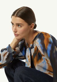 Durchsichtige Bluse in mehrfarbigen Blau-, Braun- und Cremetönen. Merkmale sind lockerer Schnitt, lange Ärmel und ein Bindeband am Hals.