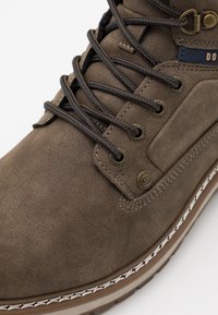Botte en cuir brun texturé avec des lacets foncés, œillets renforcés et couture blanche contrastante sur la semelle. Présente un détail de logo sur le côté.