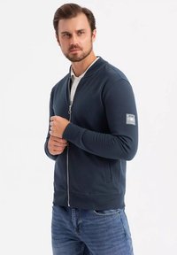 Felpa blu marina con zip, con polsini e orlo a coste, tessuto leggero, presenta un distintivo sulla manica sinistra, abbinata a una camicia con colletto sotto.