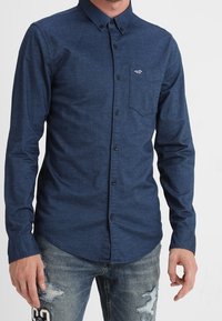 Námořnická modrá košile na knoflíky z texturované látky, s kapsou na levé hrudi a logem. V kombinaci s ošuntělými džíny.