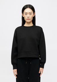 Νεαρή γυναίκα με μακριά μαύρα μαλλιά, φορώντας ένα χαλαρό μαύρο φούτερ και μαύρο παντελόνι, στέκεται μπροστά από ένα απλό αχνό φόντο.