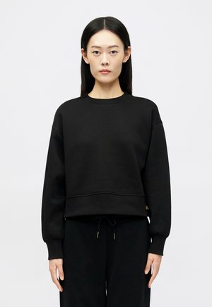 Mujer joven con cabello largo y negro, vistiendo una sudadera negra holgada y pantalones negros, de pie frente a un fondo claro y liso.