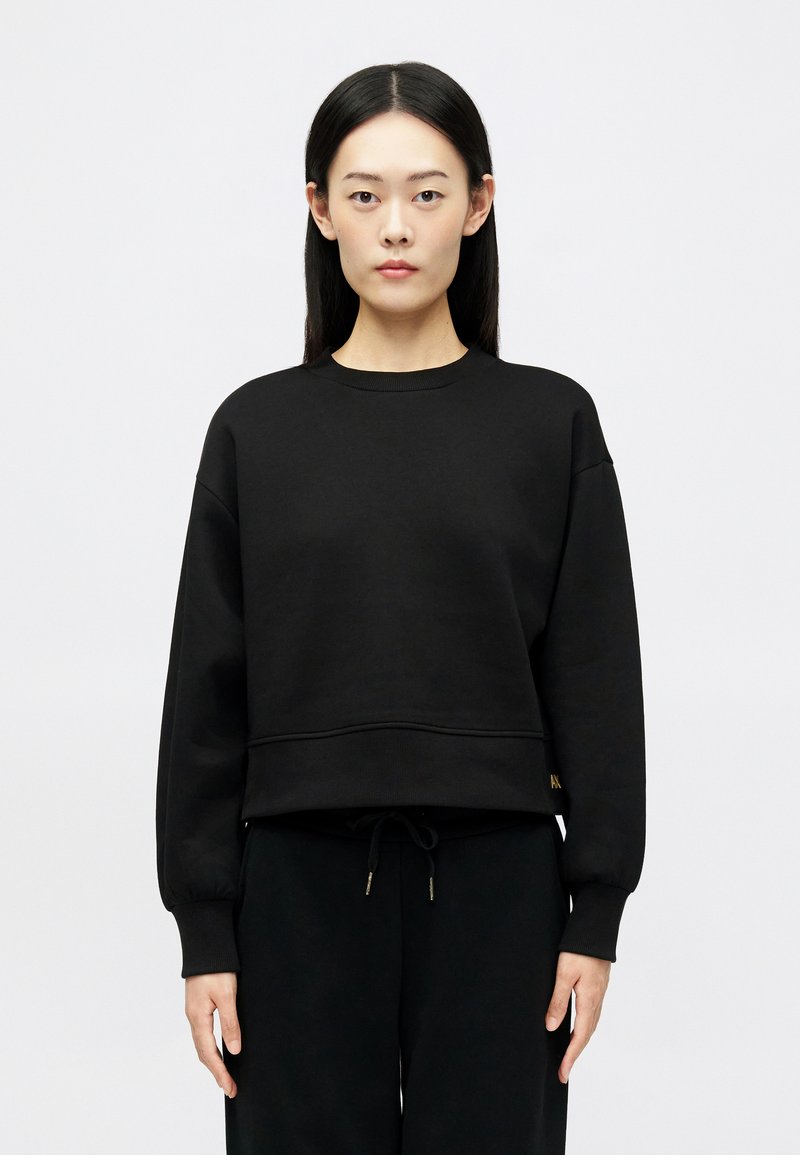 Νεαρή γυναίκα με μακριά μαύρα μαλλιά, φορώντας ένα χαλαρό μαύρο φούτερ και μαύρο παντελόνι, στέκεται μπροστά από ένα απλό αχνό φόντο.