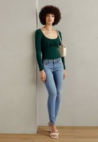 Mørkegrønn langermet topp med samlede detaljer, kombinert med lyseblå skinny jeans og hvite stroppelabber. Liten beige håndveske over skulderen.