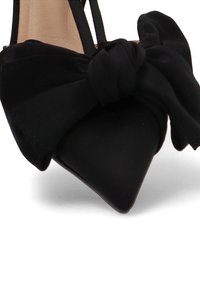 Zapatos de tacón alto negros con punta afilada, adornados con un gran lazo de tela en la parte delantera y correas en el tobillo.