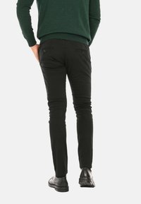 Pantaloni neri slim-fit in cotone, con tasche posteriori e una texture liscia, abbinati a scarpe nere e un maglione verde.