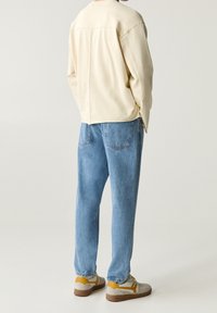 Personne portant une chemise à manches longues beige clair, un jean bleu et des baskets beige avec des accents orange, debout devant un fond clair uni.