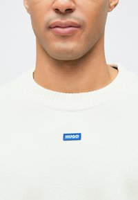 Hvit genser med rund hals som har en strikket tekstur og et blått rektangulært "HUGO" logo-lapp på venstre side av brystet.