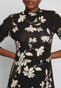 Czarna sukienka o dopasowanym kroju, zdobiona białymi kwiatowymi wzorami, z długimi rękawami i wysokim kołnierzem, wykonana z miękkiego, gładkiego materiału.