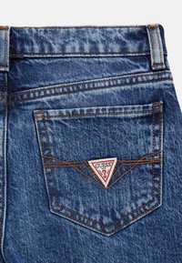 Jeans in denim con un lavaggio blu sbiadito, caratterizzati da cuciture in rame e un logo a triangolo in rosso e bianco sulla tasca posteriore.