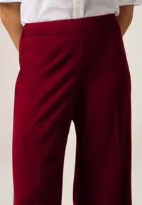Pantaloni larghi bordeaux con una texture liscia, dotati di una vita sagomata e pieghe frontali per un look classico e curato.