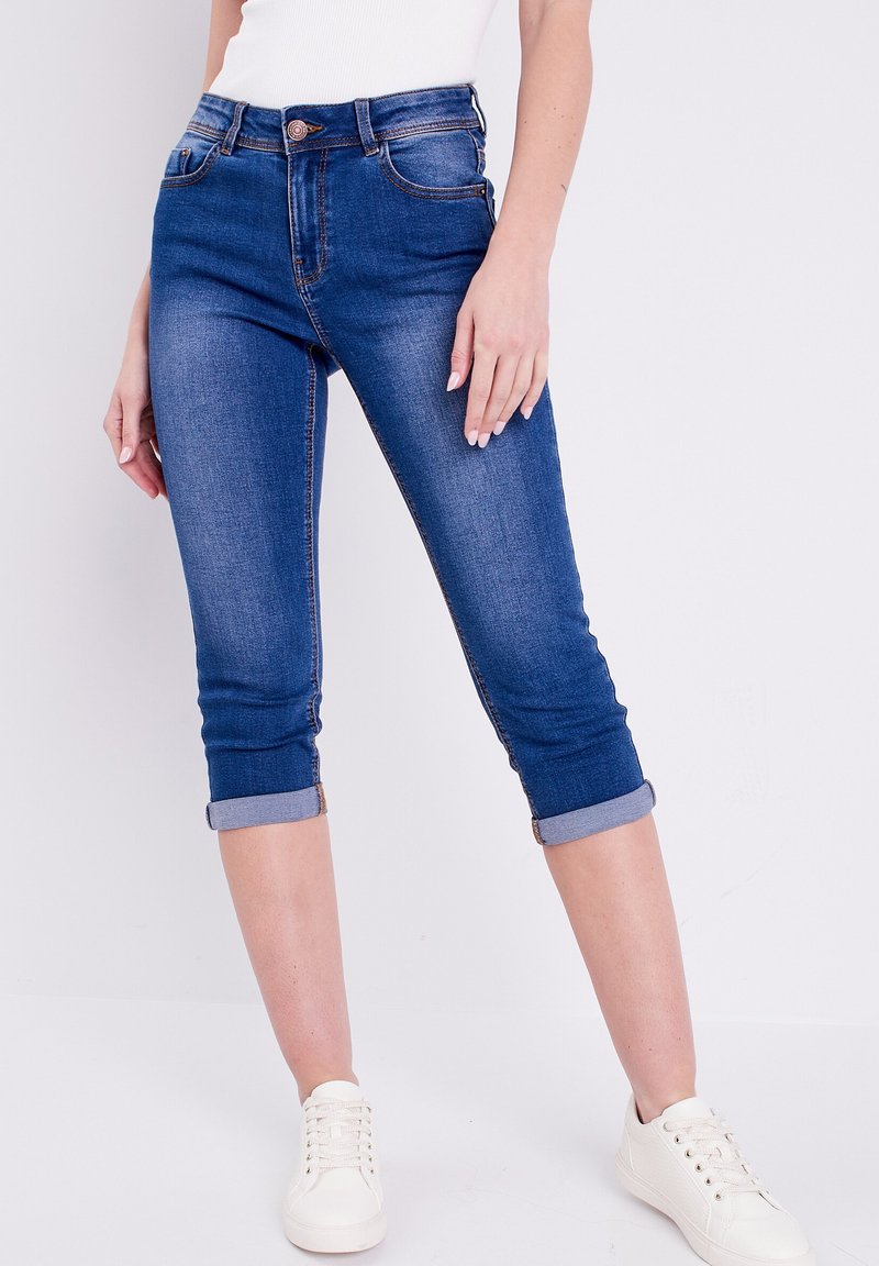 Vaqueros capri de mezclilla azul con puños enrollados, que presentan un cierre frontal con cremallera y botón, y dos bolsillos delanteros. Textura suave, lavado medio.