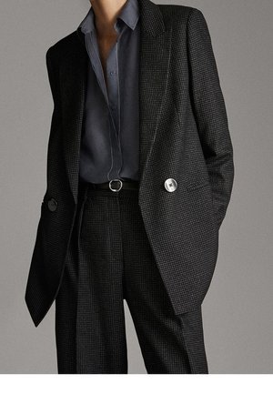 Woman wearing a dark checkered oversized blazer and matching wide-leg trousers with a dark gray button-up shirt tucked in.