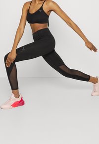 Top corto atlético negro y leggings con paneles de malla, con un patrón texturizado. Unas zapatillas deportivas en rosa y blanco completan el conjunto.