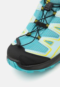 Terrengløpesko med mesh-overdel i akvablått og gule detaljer. Har en svart gummitåhette og integrert snøresystem for tilpasning.