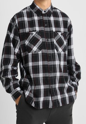 Zwart-wit-rood geruit overhemd met lange mouwen en knopen, met twee borstzakken, losjes ingestopt in een donkere broek.