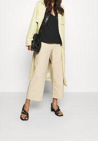 Cappotto lungo giallo chiaro, camicia in pelle nera, canotta nera, pantaloni beige a gamba larga e sandali neri con tacco a blocco e cinturini incrociati.