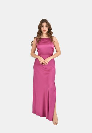 Donna con capelli lunghi e ondulati che indossa un abito in raso magenta lungo fino a terra con uno spacco, in piedi con le mani intrecciate davanti, tacchi beige visibili.