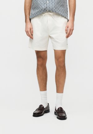 Hombre vistiendo pantalones cortos blancos con cordón, camisa de manga corta con estampado, calcetines blancos y mocasines de cuero marrón oscuro sobre un fondo liso.