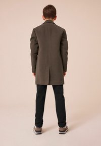 Abrigo de lana verde oliva con un diseño recto, abertura trasera y pantalones negros. Zapatillas grises y beige completan el conjunto, enfatizando un estilo casual.