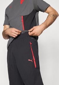Grå og sort træningsskjorte med rød lynlåsdetalje, parret med sorte bukser med røde accenter og Puma-logo. Glat stof, tætsiddende design.
